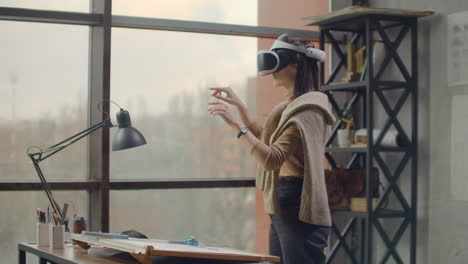 Modern-Woman-architect-in-the-office-with-large-windows-stands-in-a-virtual-reality-helmet-uses-gestures-to-manage-the-project-without-leaving-the-office.-Construction-control.-Design-project-of-the-building-and-interior